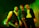 Three individuals stand in a room with green ambient lighting, each holding a musical instrument. The person on the left is holding an electric guitar and wearing a vest over a white shirt. The person in the middle is wearing a leopard print long-sleeve top and holding a guitar, while the person on the right is wearing an open jacket and a hat.