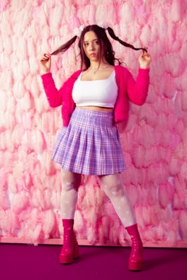 A person stands against a feather-textured pink wall, holding pigtails up on either side of the head. They are wearing a white crop top, a fluffy pink cardigan, a purple plaid skirt, white mesh tights, and pink platform boots. The overall style is playful and colorful.