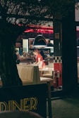 A bustling diner with retro decor, featuring checkered patterns and bright red accents. A person is seen clearing a table, wearing a red uniform. The interior is dimly lit with red neon lights and a cozy ambiance. A tree partially obscures the view from the foreground.