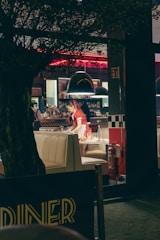 A bustling diner with retro decor, featuring checkered patterns and bright red accents. A person is seen clearing a table, wearing a red uniform. The interior is dimly lit with red neon lights and a cozy ambiance. A tree partially obscures the view from the foreground.