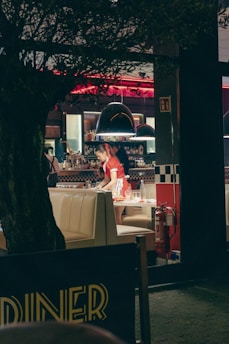 A bustling diner with retro decor, featuring checkered patterns and bright red accents. A person is seen clearing a table, wearing a red uniform. The interior is dimly lit with red neon lights and a cozy ambiance. A tree partially obscures the view from the foreground.