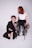 A person with curly hair and wearing a partially open black suit is kneeling on the floor. Another person with red hair is seated on a high stool, wearing a white top and a black tulle skirt. They are holding a chain attached to the neck of the person kneeling.