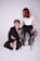 A person with curly hair and wearing a partially open black suit is kneeling on the floor. Another person with red hair is seated on a high stool, wearing a white top and a black tulle skirt. They are holding a chain attached to the neck of the person kneeling.