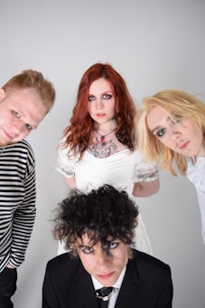 Four individuals with distinctive hairstyles and makeup pose in front of a neutral background. The person in the foreground wears a black suit, while others sport casual and vintage-inspired clothing. Vivid red, blonde, and brown hair colors stand out, and subdued expressions add intrigue.