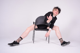 A person with curly hair is sitting on a chair backwards. They are wearing a black blazer, white shirt, and black shorts, along with fishnet socks and black shoes. The person is extending their legs widely and raising one hand with a middle finger gesture. The shoes have the words 'WE ARE NOT' inscribed on the side.