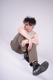 A person with a distinctive hairstyle sits on the floor, wearing a sleeveless top, wide-legged pants, and sneakers. They have a casual pose with one leg bent and the other extended, displaying fishnet socks. The sneakers have a visible message on the sole. Accessories include a silver chain necklace and a ring.