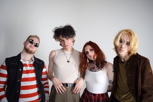 Four individuals standing against a white background, each displaying unique fashion styles. One wears a red and white striped shirt with sunglasses and a denim vest. Another has curly hair, wearing a sleeveless top and multiple necklaces. The third person has long red hair, tattoos, and is dressed in a white tank top and a red plaid skirt. The last individual has blonde hair and wears a brown jacket over an olive green shirt.