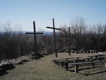 Three wooden crosses stand on a grassy area with a backdrop of leafless trees. Several wooden benches are arranged in front of the crosses, resembling an outdoor assembly or gathering space. The sky is clear and blue, suggesting a calm, open-air setting.
