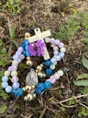 A rosary with multicolored beads lies on the ground among grass and small plants. A small crucifix and a medallion with a religious figure are attached to the beads. A cluster of purple flowers is interwoven with the rosary.