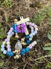 A rosary with multicolored beads lies on the ground among grass and small plants. A small crucifix and a medallion with a religious figure are attached to the beads. A cluster of purple flowers is interwoven with the rosary.