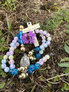 A rosary with multicolored beads lies on the ground among grass and small plants. A small crucifix and a medallion with a religious figure are attached to the beads. A cluster of purple flowers is interwoven with the rosary.