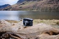 A black camera rests on a large, weathered piece of driftwood with a serene lake and rolling hills in the background. The landscape features calm water, grassy areas, and mountainous terrain partially covered with trees and shrubs.
