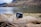 A black camera rests on a large, weathered piece of driftwood with a serene lake and rolling hills in the background. The landscape features calm water, grassy areas, and mountainous terrain partially covered with trees and shrubs.
