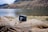 A black camera rests on a large, weathered piece of driftwood with a serene lake and rolling hills in the background. The landscape features calm water, grassy areas, and mountainous terrain partially covered with trees and shrubs.