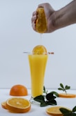 A hand squeezes an orange over a tall glass filled with orange juice. Slices of fresh oranges and green leaves are arranged around the glass, creating a refreshing and vibrant setup.