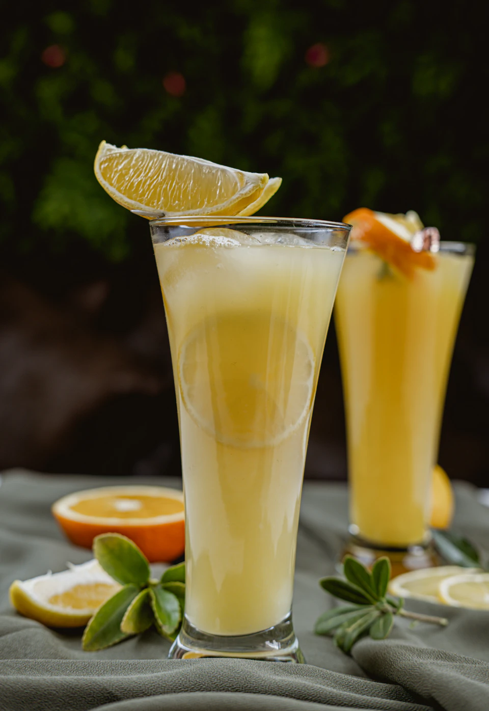 The image presents a vibrant scene set on a table draped with a green tablecloth, bathed in soft light that accentuates its details. Two tall glasses filled with a yellow liquid are placed at opposite ends of the table, each adorned with slices of orange and lemon. The glass on the left is almost empty, while the one on the right appears to be brimming with the yellow liquid. A slice of orange rests near the bottom of the image, adding a pop of color against the green backdrop.