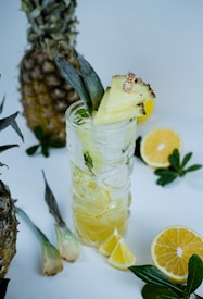 A tall glass filled with a yellow beverage garnished with pineapple slices and a small decorative pineapple. Surrounded by whole pineapples, lemon slices, and green leaves, creating a tropical and refreshing atmosphere.