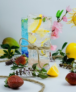 Two tall, intricately designed glasses are filled with a refreshing lemon and herb-infused drink, with slices of lemon and mint leaves inside. One glass is adorned with a rustic twine bow. Surrounding the glasses are fresh strawberries, lemon slices, and decorative flowers and leaves, creating a vibrant and natural display.