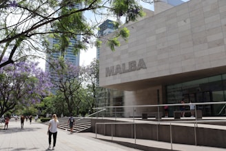 A modern building with a large sign reading 'MALBA' is situated near a walkway lined with trees that have purple blossoms. People are walking along the path, which is bordered by lush green trees and a few tall buildings in the background.