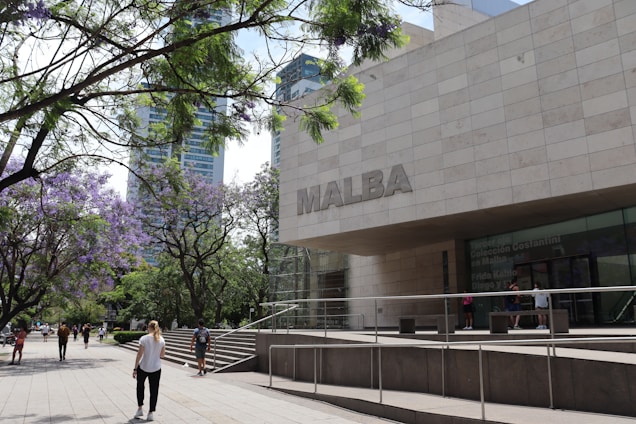 A modern building with a large sign reading 'MALBA' is situated near a walkway lined with trees that have purple blossoms. People are walking along the path, which is bordered by lush green trees and a few tall buildings in the background.