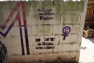 The image features a concrete wall with graffiti. There is text in Spanish highlighting feminist themes, including phrases such as 'No somos histéricas, somos históricas.' A purple feminist symbol with a raised fist is also visible. The wall has a greenish hue due to moss or staining, and there are some colored geometric shapes in purple and red on the left side.