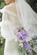 A bride in a lace wedding dress is holding a bouquet of various flowers, including purple and pink blooms with green leaves. The dress features an off-the-shoulder design with a veil cascading down her back. The background is blurred greenery.