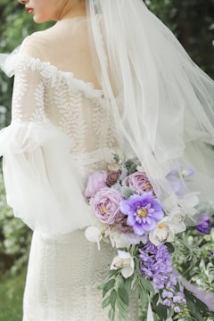 A bride in a lace wedding dress is holding a bouquet of various flowers, including purple and pink blooms with green leaves. The dress features an off-the-shoulder design with a veil cascading down her back. The background is blurred greenery.