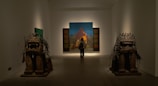 A person stands in an art gallery, observing a large painting in front of them. The painting depicts a mountainous landscape with distinct colors. On either side of the room, there are two large, detailed sculptures resembling stylized animals.