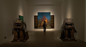 A person stands in an art gallery, observing a large painting in front of them. The painting depicts a mountainous landscape with distinct colors. On either side of the room, there are two large, detailed sculptures resembling stylized animals.