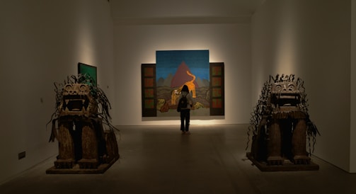 A person stands in an art gallery, observing a large painting in front of them. The painting depicts a mountainous landscape with distinct colors. On either side of the room, there are two large, detailed sculptures resembling stylized animals.