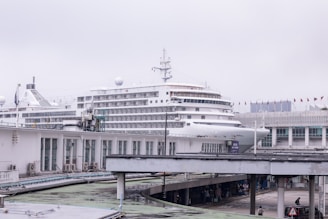 A large cruise ship docked at a port, with several decks visible, towering over nearby structures. The background includes buildings with multiple windows and a row of small flags. Below, there are walkways with people walking, suggesting a busy port area.