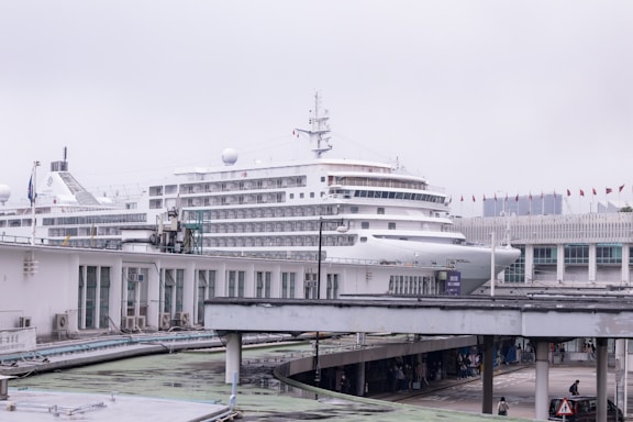 A large cruise ship docked at a port, with several decks visible, towering over nearby structures. The background includes buildings with multiple windows and a row of small flags. Below, there are walkways with people walking, suggesting a busy port area.
