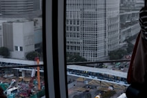 The image depicts a cityscape with a focus on a large building characterized by a grid-like pattern of windows. A construction site is visible below, featuring cranes, construction vehicles, and workers. In the foreground, there is a metal structure, possibly part of a window frame. The sky is overcast, giving the scene a somewhat gloomy atmosphere.