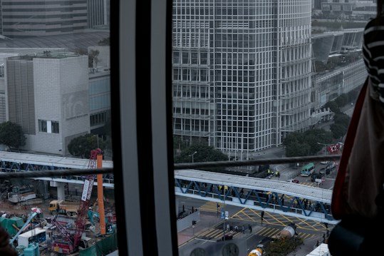 The image depicts a cityscape with a focus on a large building characterized by a grid-like pattern of windows. A construction site is visible below, featuring cranes, construction vehicles, and workers. In the foreground, there is a metal structure, possibly part of a window frame. The sky is overcast, giving the scene a somewhat gloomy atmosphere.