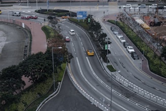 The image captures an aerial view of a multi-lane urban road with cars traveling in both directions. The central road is curved and flanked by greenery and trees. A row of parked cars lines one side, while construction work appears to be taking place on the opposite side, with visible equipment and fencing. Sidewalks for pedestrians border the roads, and several traffic signs are present.