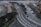 The image captures an aerial view of a multi-lane urban road with cars traveling in both directions. The central road is curved and flanked by greenery and trees. A row of parked cars lines one side, while construction work appears to be taking place on the opposite side, with visible equipment and fencing. Sidewalks for pedestrians border the roads, and several traffic signs are present.