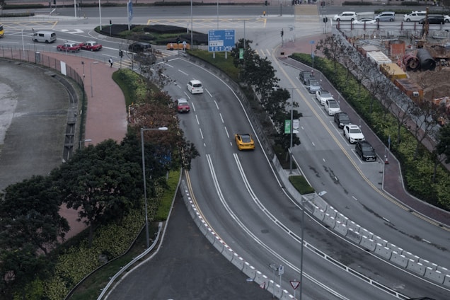 The image captures an aerial view of a multi-lane urban road with cars traveling in both directions. The central road is curved and flanked by greenery and trees. A row of parked cars lines one side, while construction work appears to be taking place on the opposite side, with visible equipment and fencing. Sidewalks for pedestrians border the roads, and several traffic signs are present.