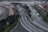 The image captures an aerial view of a multi-lane urban road with cars traveling in both directions. The central road is curved and flanked by greenery and trees. A row of parked cars lines one side, while construction work appears to be taking place on the opposite side, with visible equipment and fencing. Sidewalks for pedestrians border the roads, and several traffic signs are present.