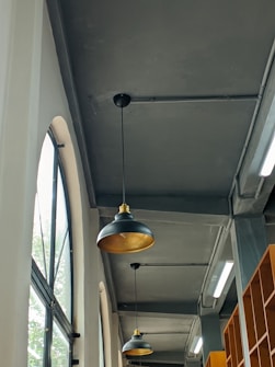 The image depicts an indoor setting with a high ceiling and industrial design. There are large arched windows on the left side allowing natural light to come in. Two black pendant lights with gold interiors are hanging from the ceiling. Shelving units are visible on the right side of the image. The color scheme is predominantly gray with hints of amber from the lights and wooden shelves.