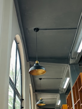 The image depicts an indoor setting with a high ceiling and industrial design. There are large arched windows on the left side allowing natural light to come in. Two black pendant lights with gold interiors are hanging from the ceiling. Shelving units are visible on the right side of the image. The color scheme is predominantly gray with hints of amber from the lights and wooden shelves.
