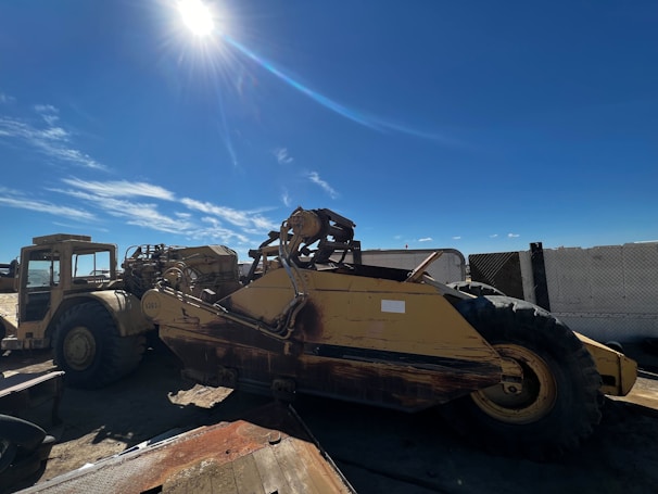A large, heavy construction vehicle sits on a dirt lot under a bright blue sky. The sunlight casts stark shadows, emphasizing the rugged appearance of the machinery. The vehicle appears weathered with visible rust on its metal surface.
