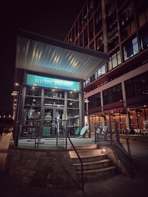 A corner donut shop with a modern design, featuring a prominent turquoise sign that reads 'District Doughnut'. The shop entrance is elevated by stone steps and surrounded by a metal railing. The adjacent building has large glass windows illuminated by warm indoor lighting.