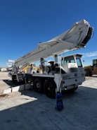 A large mobile crane is parked on a concrete surface in an industrial area. The crane's boom is extended upwards towards the clear blue sky. It is mounted on a white truck with six wheels, and there are various components visible, such as hydraulic cylinders and cables. Nearby, additional machinery and equipment can be seen, suggesting a construction or industrial setting.