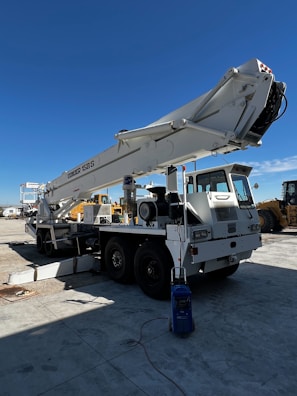A large mobile crane is parked on a concrete surface in an industrial area. The crane's boom is extended upwards towards the clear blue sky. It is mounted on a white truck with six wheels, and there are various components visible, such as hydraulic cylinders and cables. Nearby, additional machinery and equipment can be seen, suggesting a construction or industrial setting.
