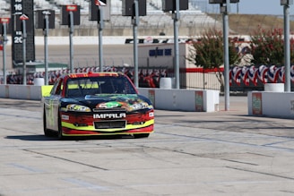 A brightly colored race car with the word 'Impala' on the front bumper is parked on a tarmac with a series of tall poles in the background. There are race track markings on the ground, and the surroundings include a fenced area decorated with red, white, and blue banners.