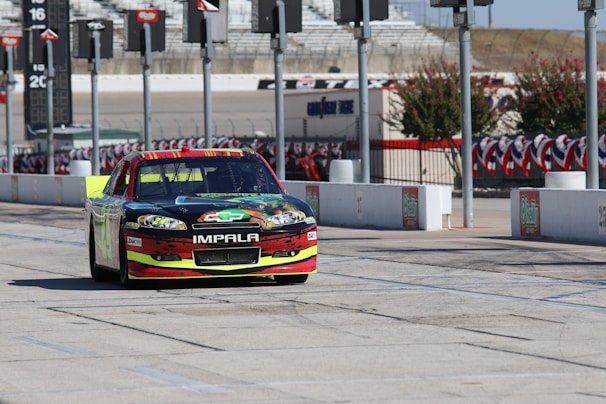 A brightly colored race car with the word 'Impala' on the front bumper is parked on a tarmac with a series of tall poles in the background. There are race track markings on the ground, and the surroundings include a fenced area decorated with red, white, and blue banners.