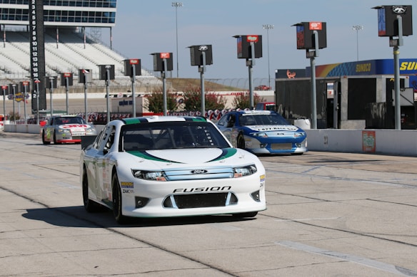 Three race cars are on a racetrack near a series of pit stalls. The foreground features a white Ford Fusion car, while two other cars are visible behind it. There are checkered flag designs and corporate logos along the pit lane. The background shows grandstands and a large scoreboard.
