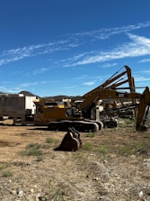 Several large construction vehicles, including excavators and machinery, are parked on a dirt area with sparse grass. The sky above is clear and blue with light clouds.
