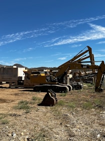 Several large construction vehicles, including excavators and machinery, are parked on a dirt area with sparse grass. The sky above is clear and blue with light clouds.