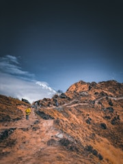 A group of hikers treks along a rugged mountain path under a vast, cloudy sky. The terrain is rocky and barren, with patches of grass and sparse vegetation. Snow-capped peaks are visible in the distance, adding a dramatic backdrop.